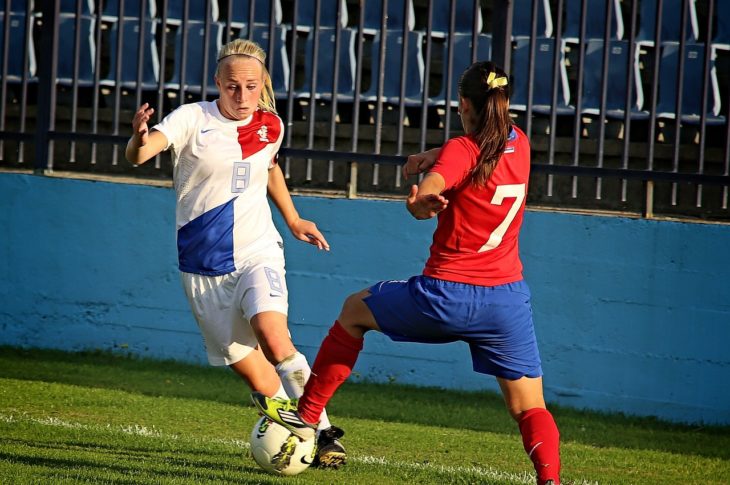 Zwei Fußballspielerinnen bei einem Spiel. Eine trägt ein weißes Trikot, die andere ein rotes