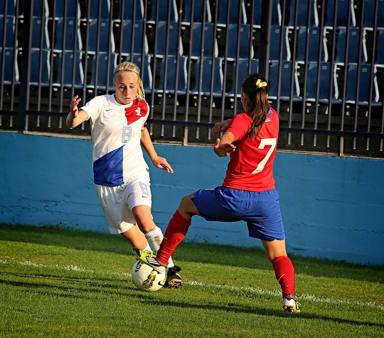 Zwei Fußballspielerinnen bei einem Spiel. Eine trägt ein weißes Trikot, die andere ein rotes